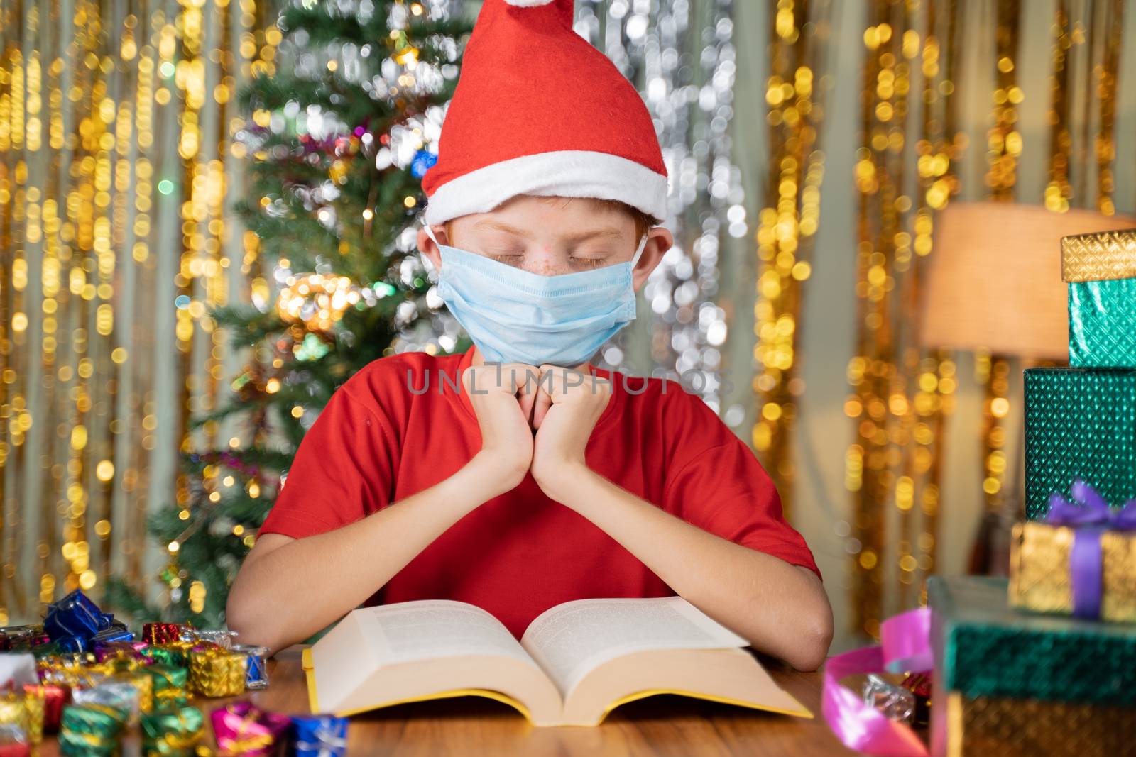 Kid with red Santa hat praying in front of Bible on Christmas decorated background with gifts on table - Concept of prayer for a Christmas eve