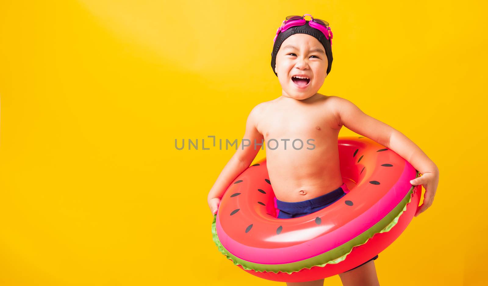 Summer vacation concept, Portrait Asian happy cute little child boy wear goggles and swimsuit hold watermelon inflatable ring, Kid having fun on summer vacation, studio shot isolated yellow background