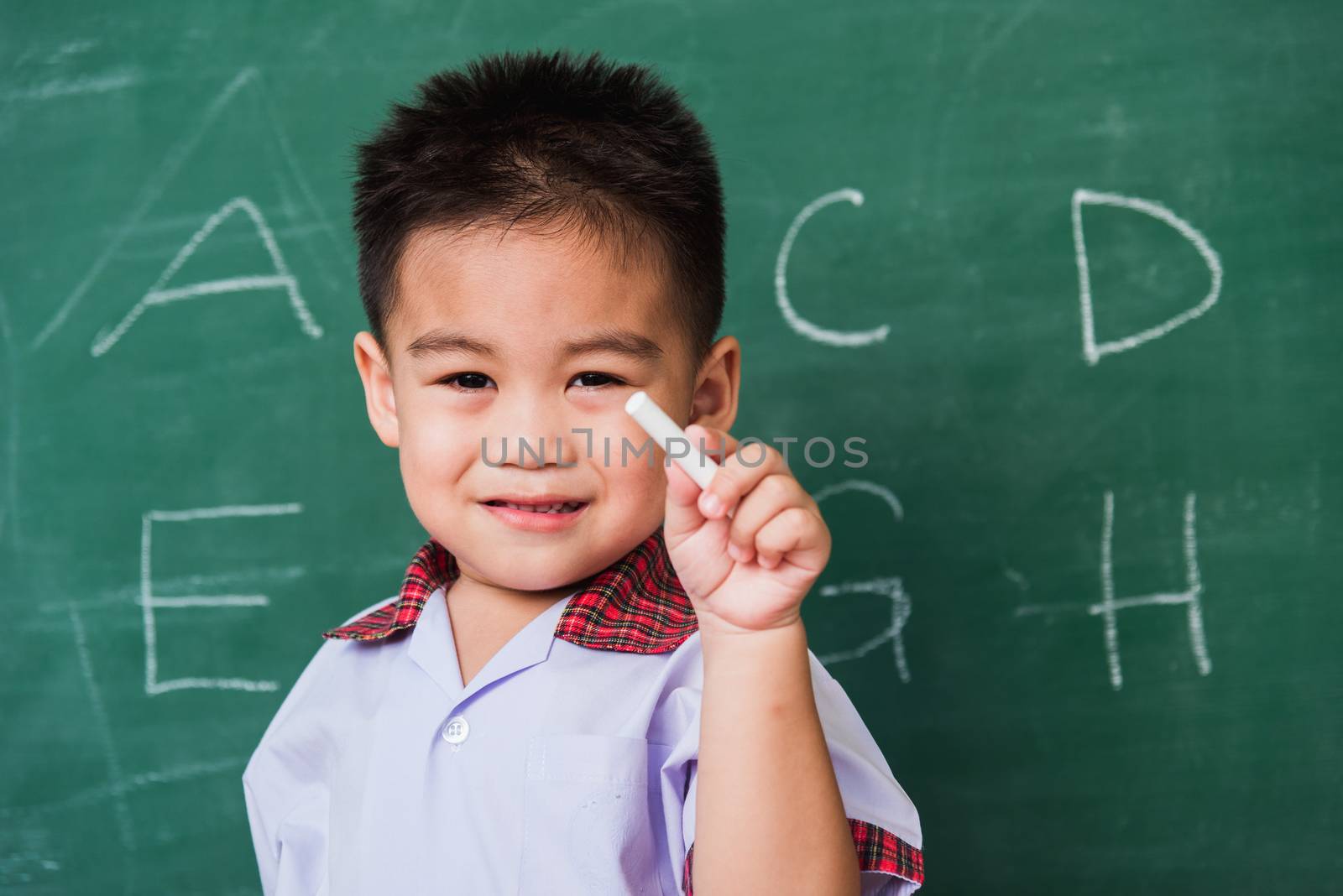Back to School. Happy Asian funny cute little child boy kindergarten preschool smile in student uniform hold white chalk after write ABC with on green school blackboard, First time to school education