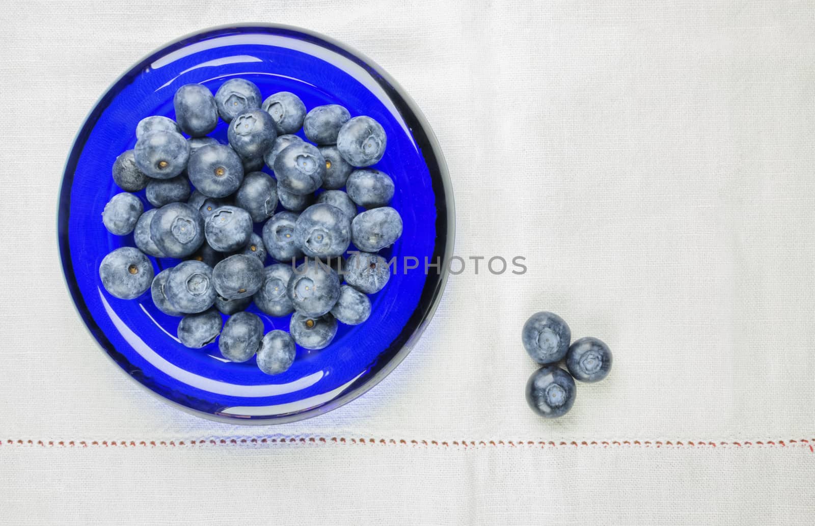 Beautiful blueberries in a blue glass bowl ,the bowl is on a white cloth , three blueberries are in the right corner of the frame