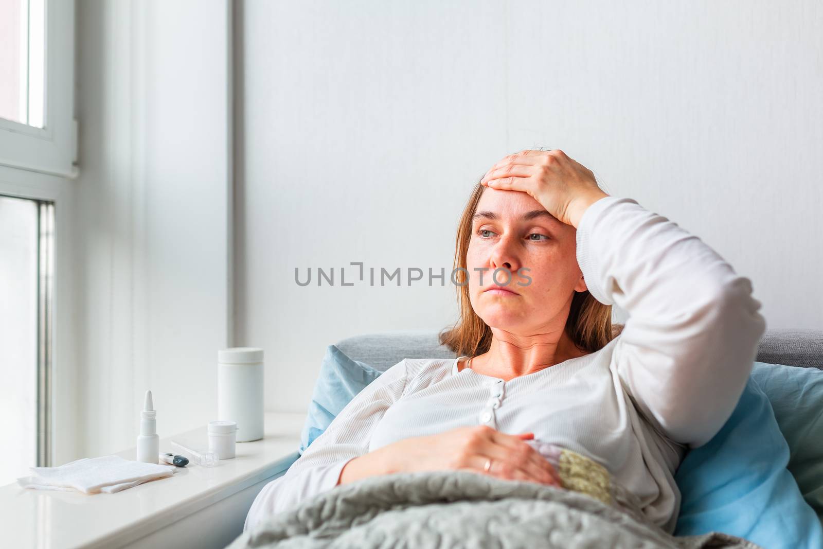 Sick woman with headache and fever lying under the blanket. Sick woman staying in bed with temperature durong coronavirus pandemic. Sick woman covered with a blanket lying in bed with high fever and a flu, resting.