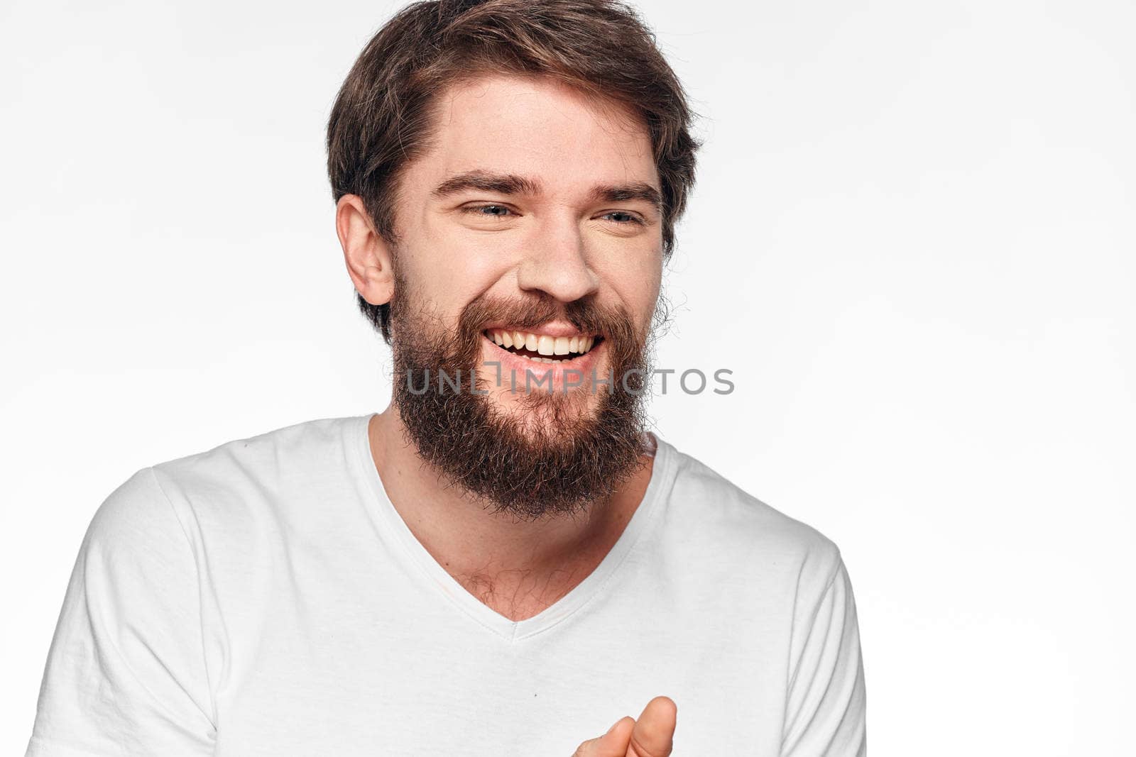 cheerful bearded man in a white t-shirt emotions gestures with his hands light background studio by SHOTPRIME