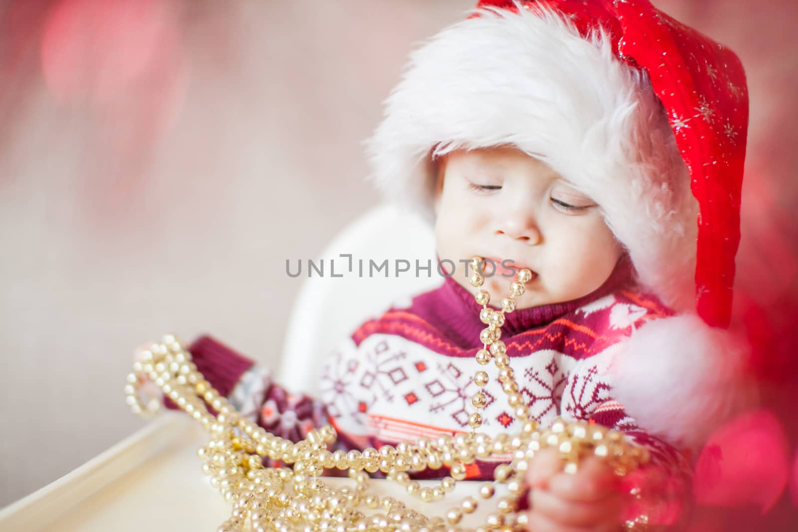 A little baby girl in a New Year's hat of Santa Claus examines and plays with New Year's decorations. Merry Christmas and Happy New Year greetings.