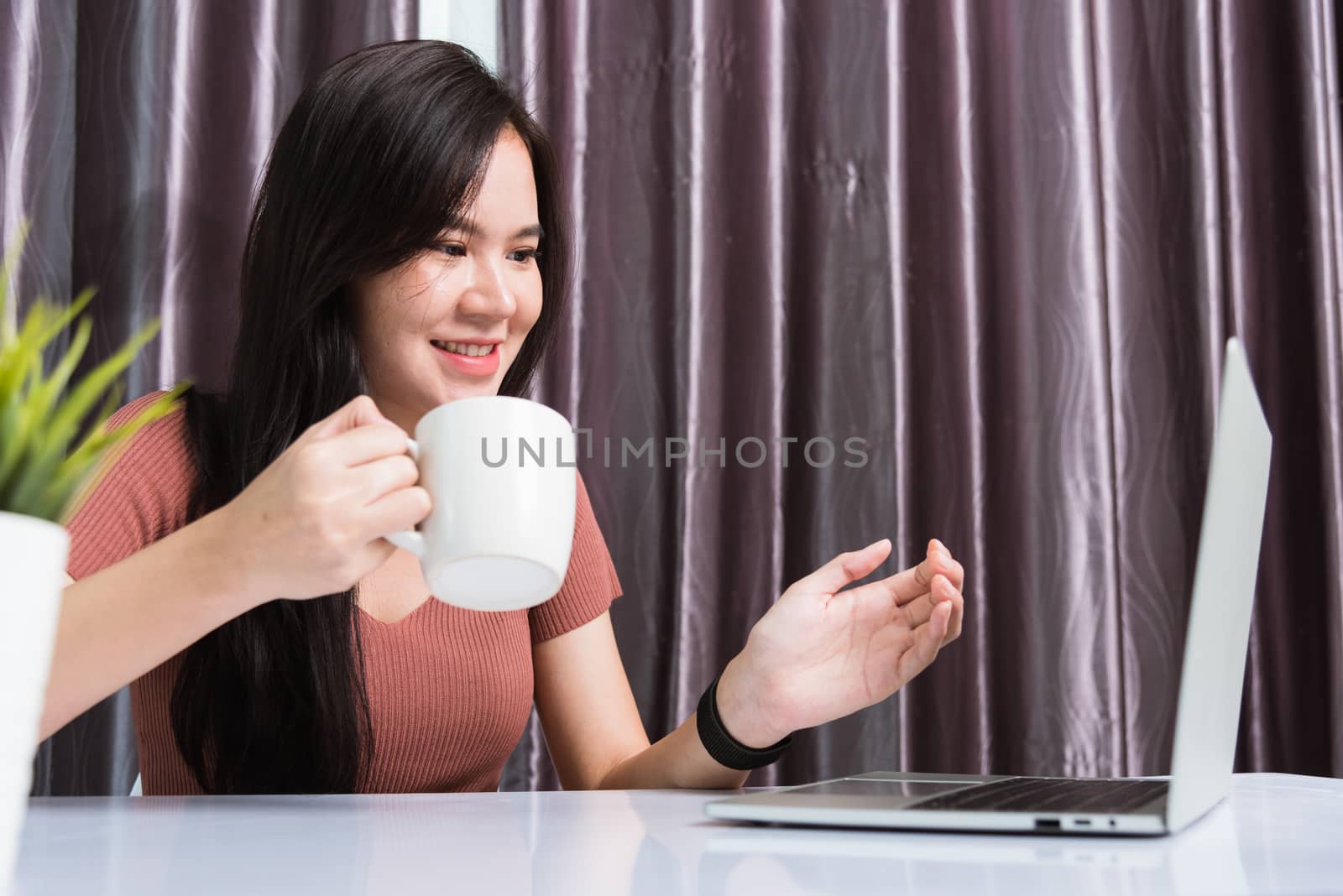 Work from home, Smiling Happy Asian business young beautiful woman sit on desk workspace video call conferencing by webcam laptop computer technology and raise your hand explain the job at home office