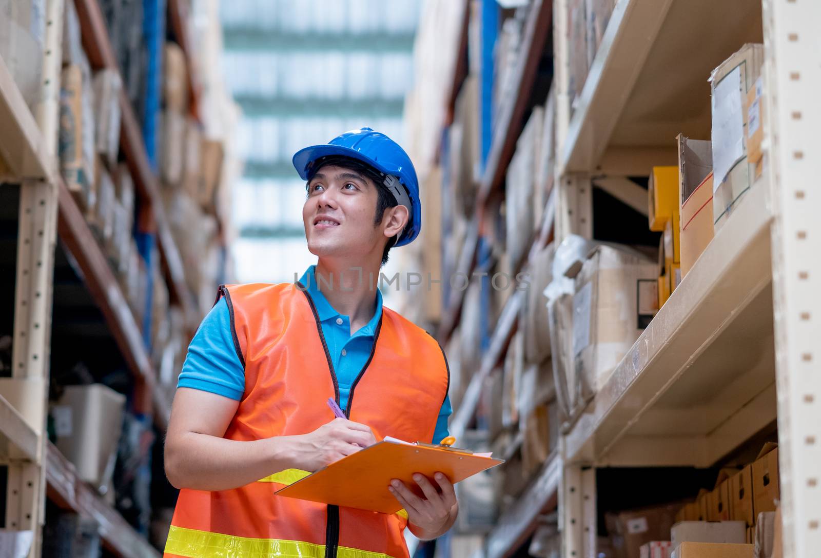 Warehouse man or factory worker with blue hard hat and uniform stand between shelves in workplace. Concept of good management and happiness of staff during work in industrial business.