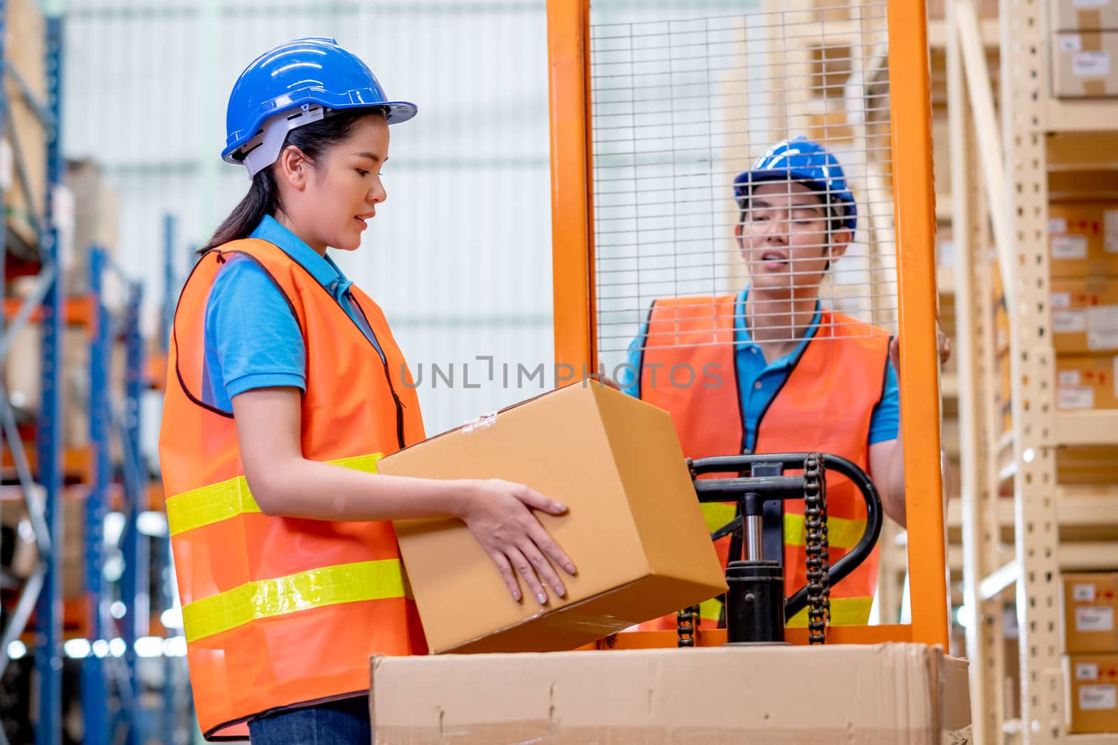 Warehouse woman or factory worker hold package box in front of hand stacker or hydraulic cart that control by her co-worker man in workplace area for delivery industrial business.