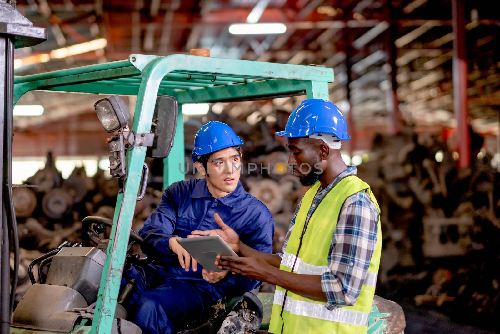 Asian technician or factory worker sit on forklift discuss about work using tablet with his co-worker African American man in automotive workplace area during day time for good industrial teamwork.