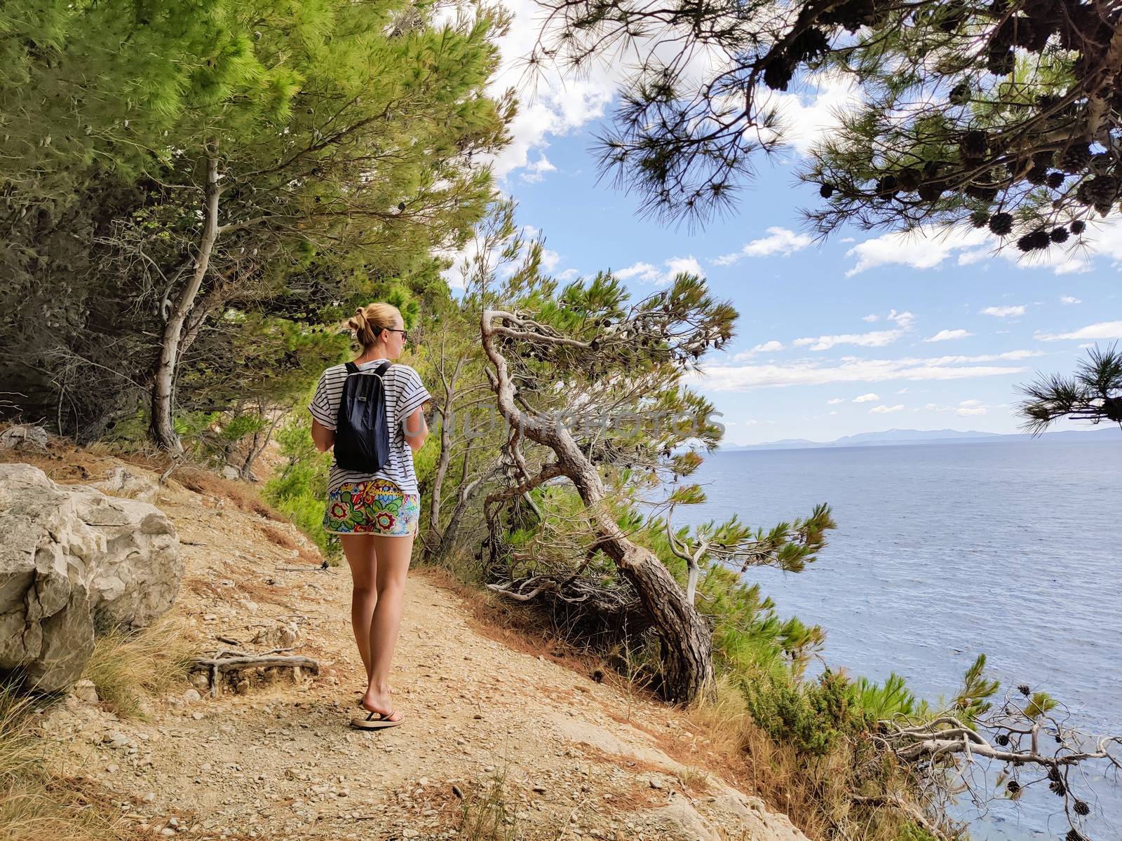 Young active feamle tourist wearing small backpack walking on coastal path among pine trees looking for remote cove to swim alone in peace on seaside in Croatia. Travel and adventure concept.