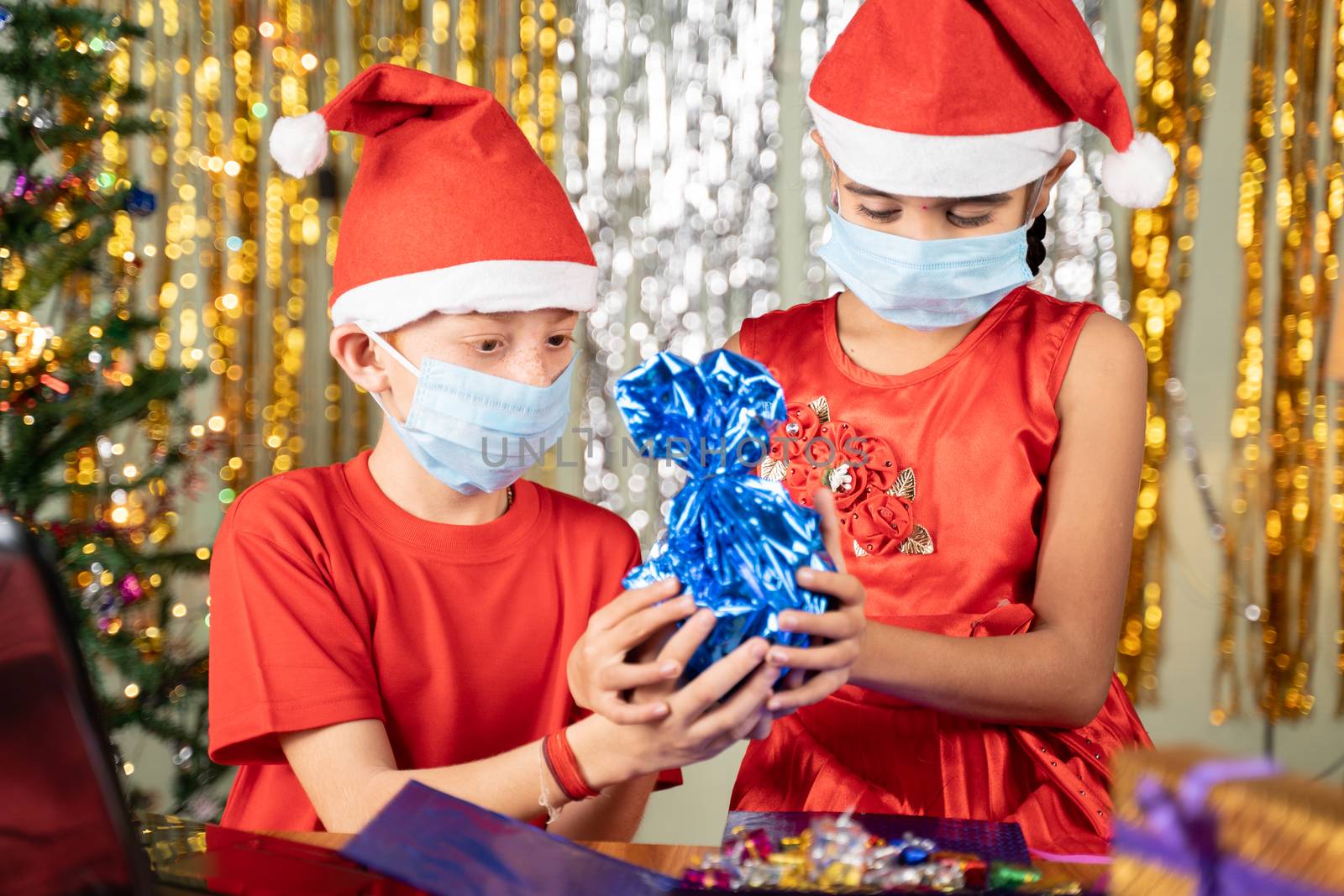 Kids in medical mask in front of laptop Opening gift at home with decorated background during Christmas eve - concept of distant xmas celebration due to coronavirus or covid-19 pandemic