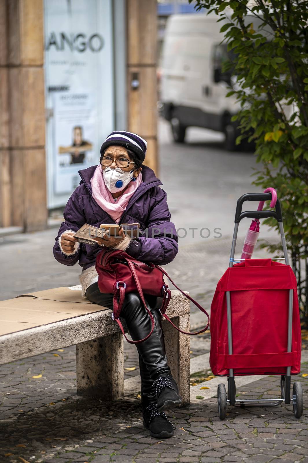 terni,itali november 05 2020:elderly oriental woman with medical mask sitting on a bench on the phone