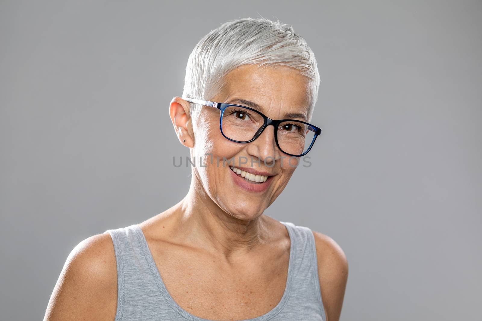 Beautiful smiling cute senior woman with short white hair and glasses posing in front of gray background