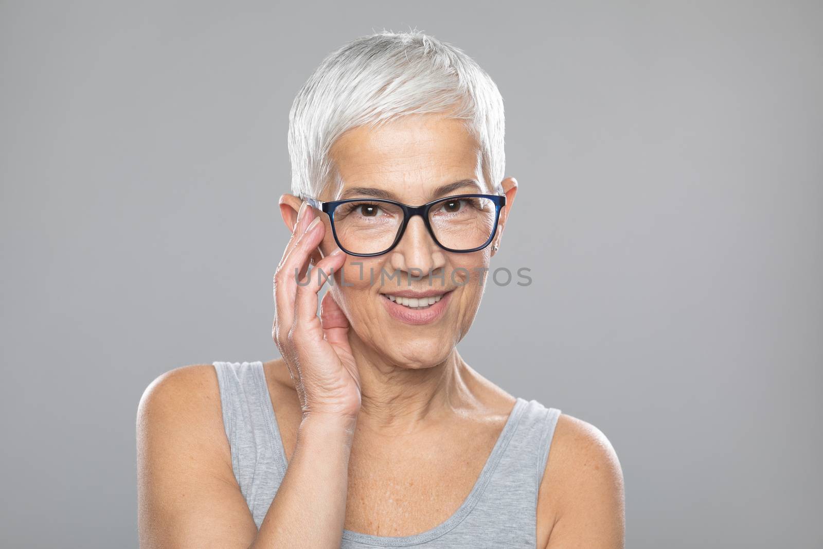Beautiful smiling cute senior woman with short white hair and glasses posing in front of gray background