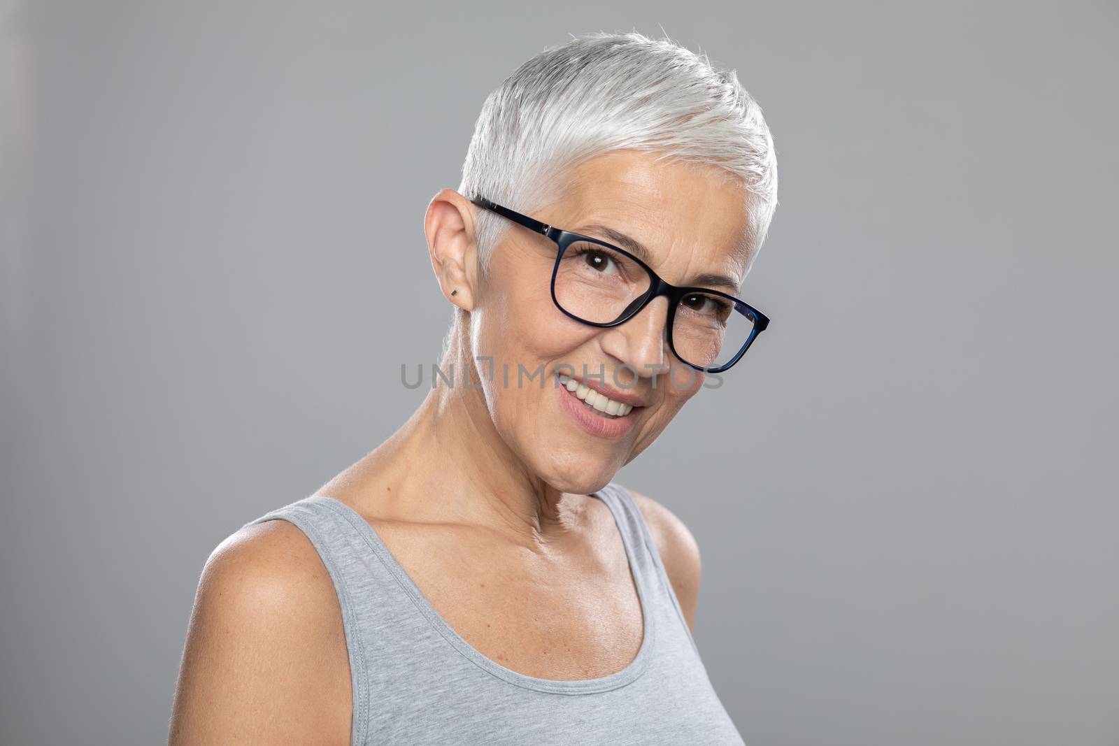 Beautiful cute smiling senior woman with short white hair and glasses posing in front of gray background