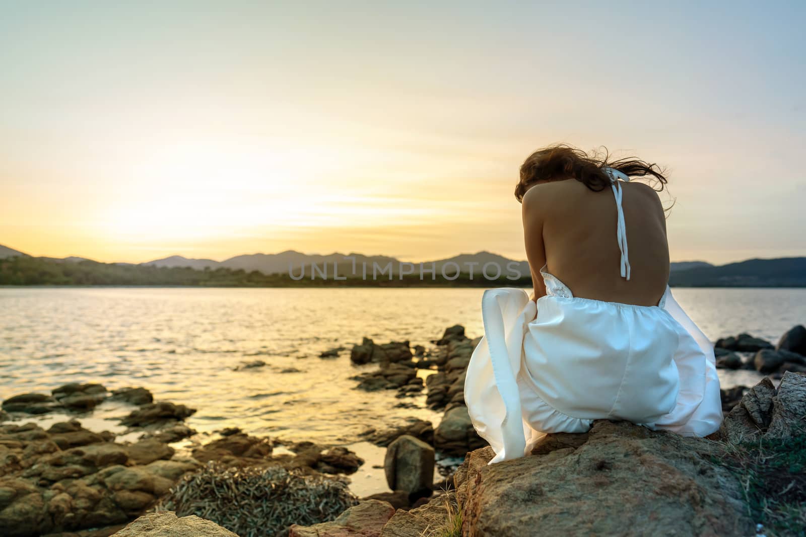 Unrecognizable pensive young caucasian woman in white wedding dress sits on the rocks by the sea at sunset with her head turned down in sadness - Concept of lost love and loneliness