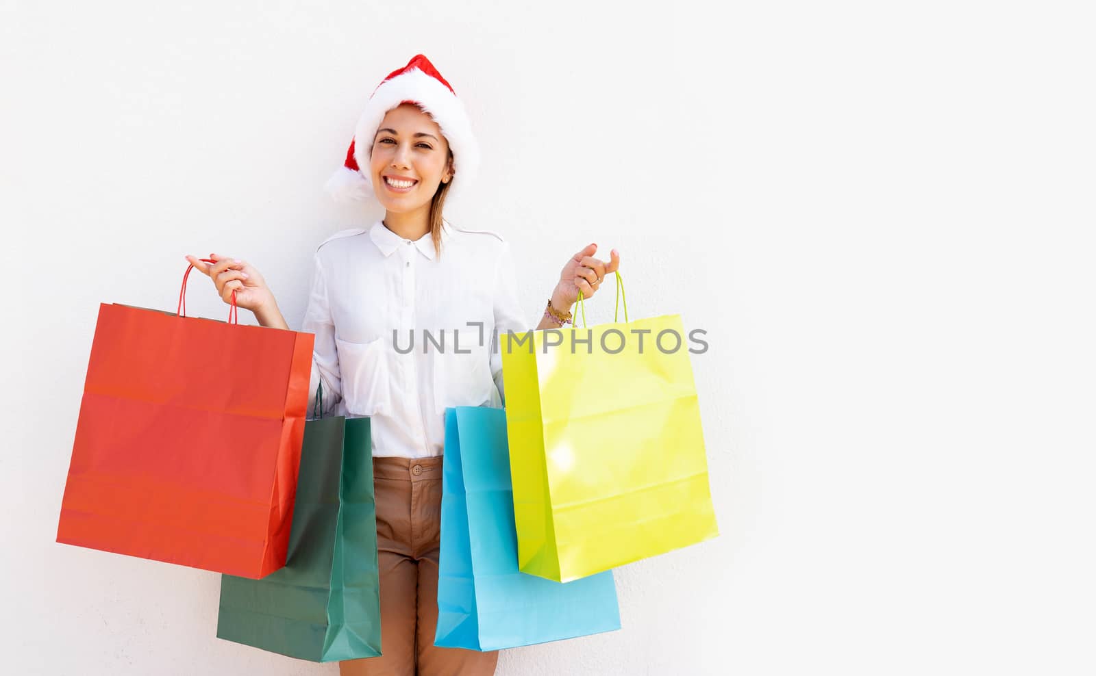 Young beautiful caucasian blonde woman standing on white background with Santa Claus hat holding many colored shopping bag and smiling looking at the camera - Buying Christmas gifts to give happiness by robbyfontanesi