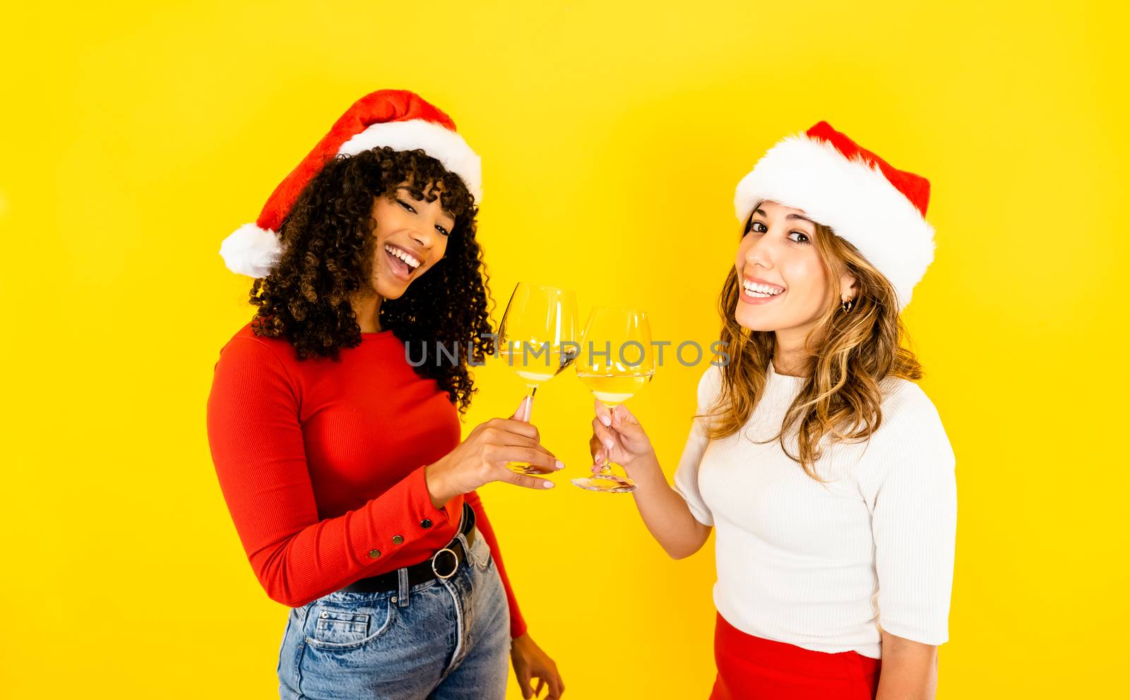 Christmas unites racial diversity - A beautiful blonde Caucasian toasts with her female black Hispanic friend wearing a Santa hat - Two isolated multiracial women model on yellow background copy space