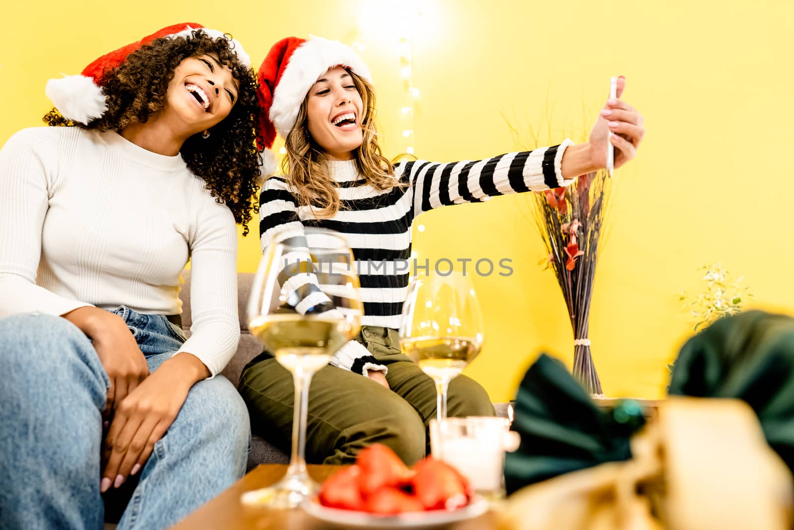 Family holidays with diversity: beautiful blonde takes a selfie at home with her black Hispanic girlfriend wearing Santa hat - Modern people different relationships and habits with technology by robbyfontanesi