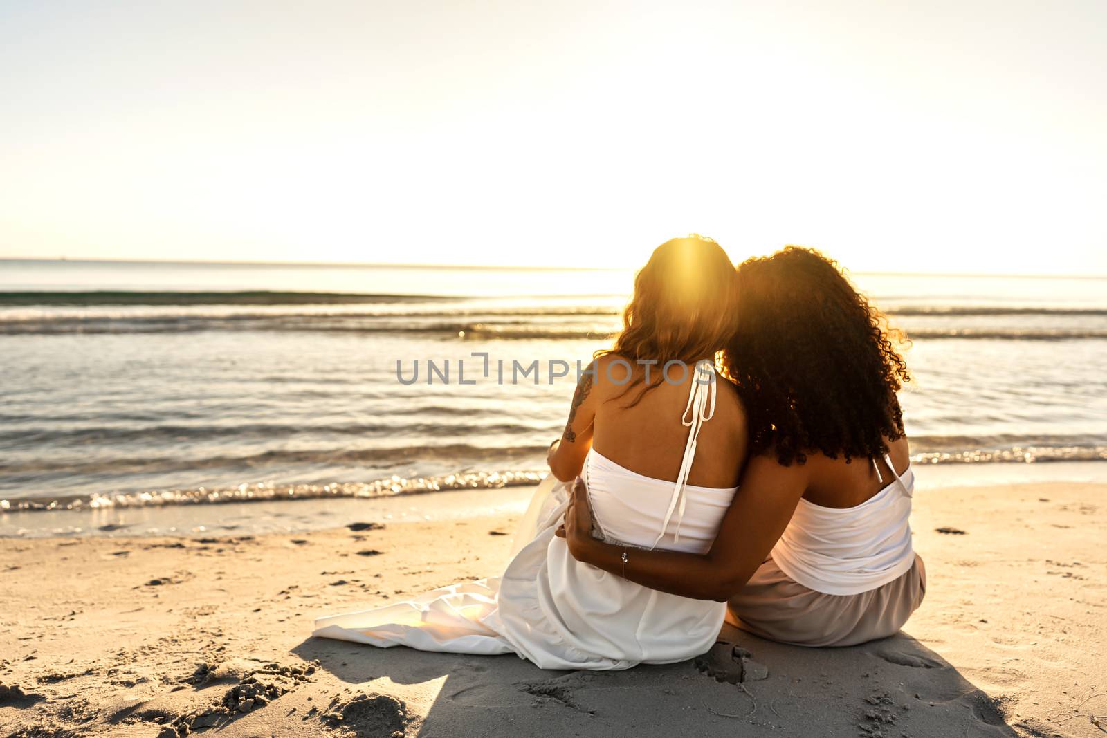 Tenderness and diversity between women who love each other - Black Hispanic curly brunette embracing her blonde tattooed girlfriend sitting on the seashore at sunset or dawn - Unrecognizable women by robbyfontanesi
