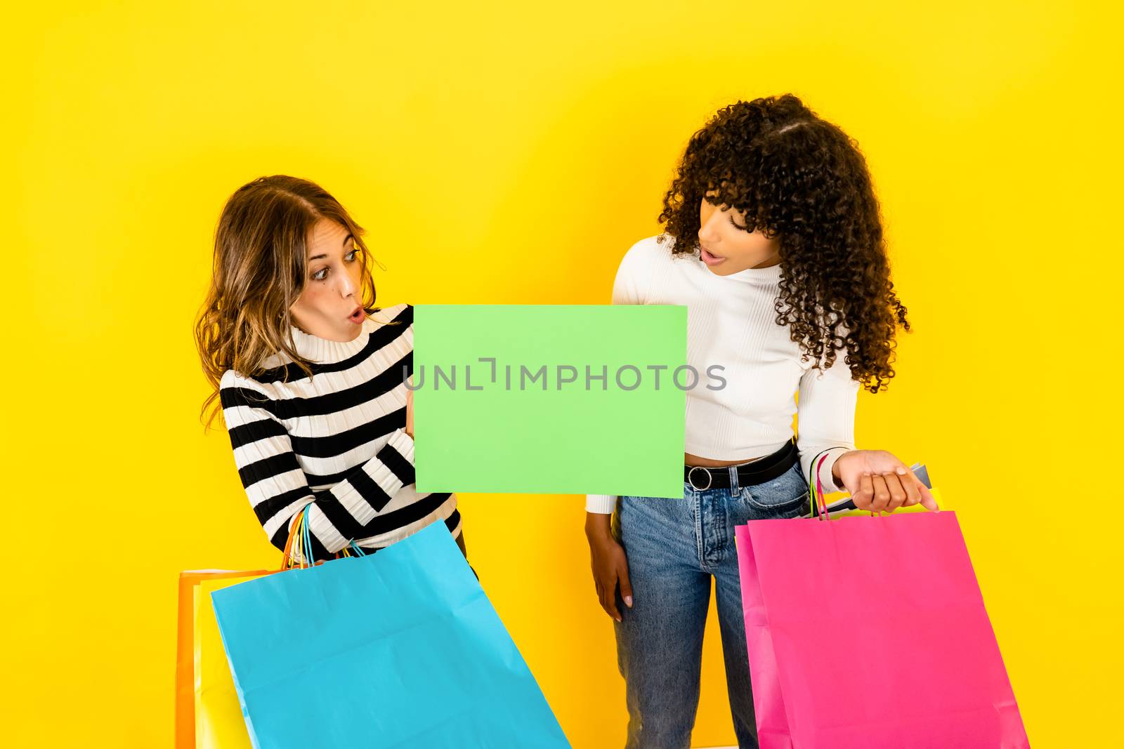 Colors and fun for shopping - Two beautiful young women look at an empty sign that I hold in my hand, ideal as a copy space - Multiracial female models excited and surprised holding some shopping bags by robbyfontanesi