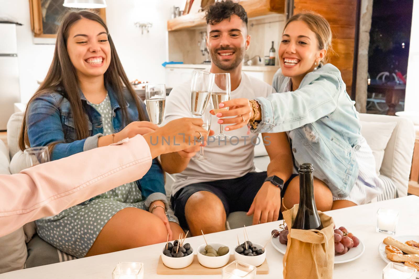 Young friends having fun toasting with champagne at home with aperitif snacks on the table - Prosecco wine to forge bonds between people that want to enjoy their life - Focus on glasses by robbyfontanesi