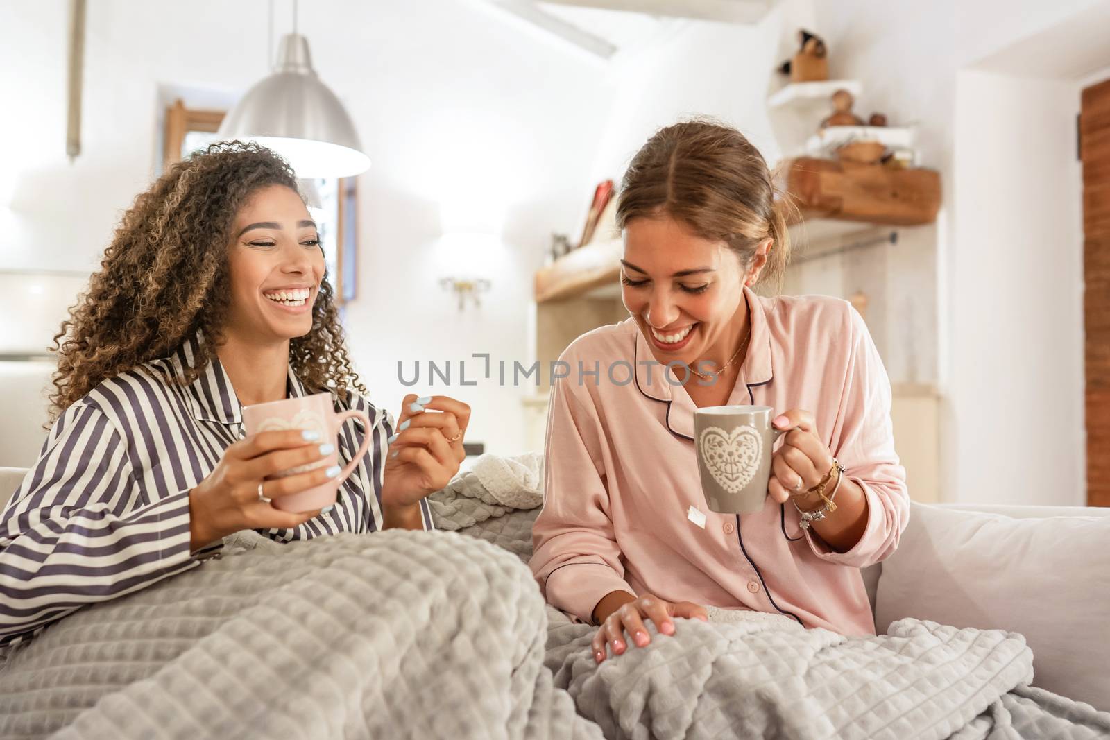 Family habits with diversity: multiracial homosexual female couple having fun at home sitting at the sofa drinking a cup of tea under a blanket - Two beautiful young women bonding each other