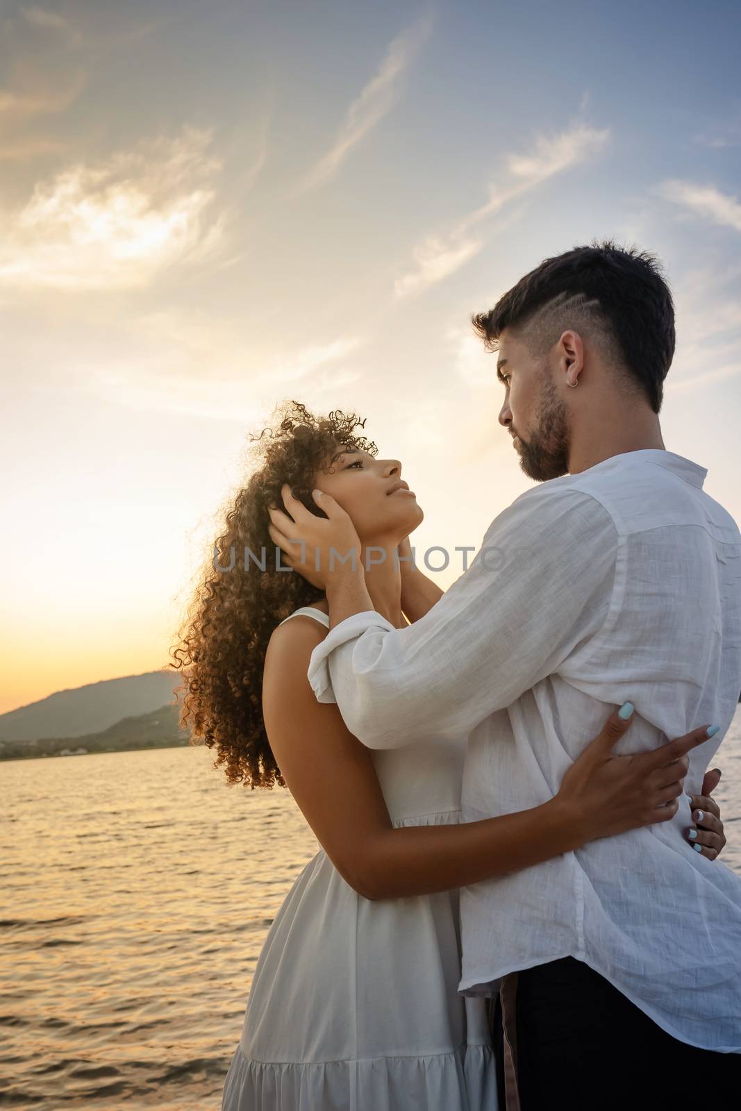 Young attractive caucasian bearded man with modern haircut holds his black Hispanic girlfriend's head in his hands looking into her eyes while she hugs him - Multiracial couple romance scene at sunset by robbyfontanesi