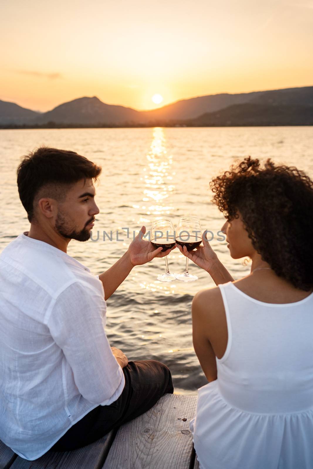 Romance scene of multiracial couple sitting on a pier at sunset or dawn toasting with red wine looking each other in the eyes - Attractive man bonding with her Hispanic girlfriend - Focus on glasses