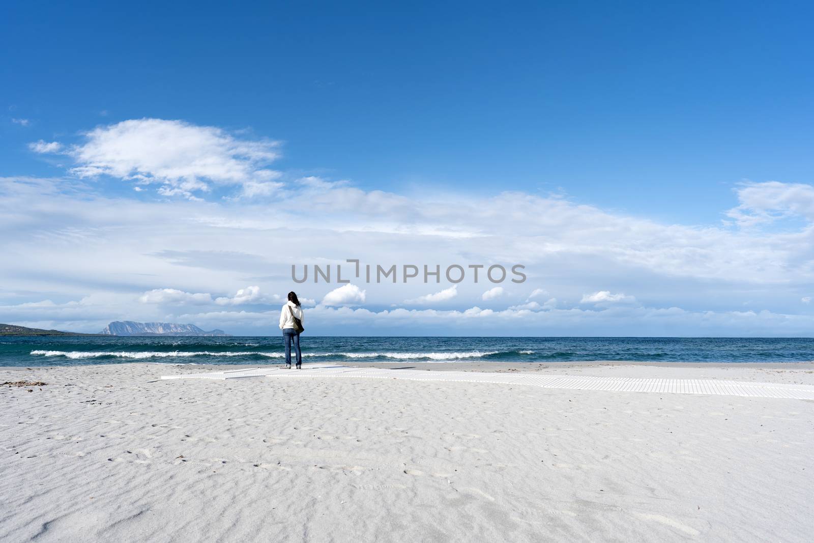 Young unrecognizable woman standing alone by the sea with cloudy sky, looks at the horizon thoughtfully - Pensive single female people in winter on the white beach with Tavolara mountain on background