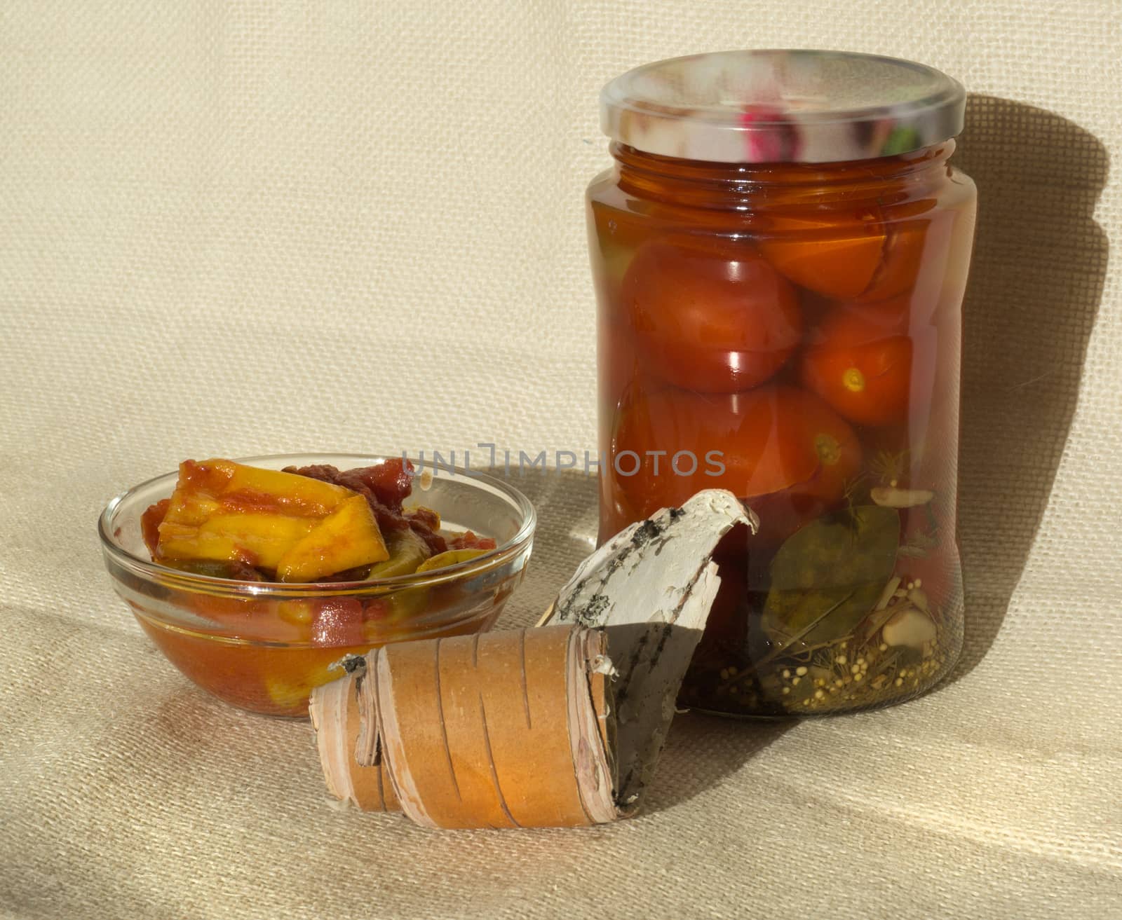 A jar of pickled tomatoes, a bowl of pickled hot peppers on a background of flax and birch bark