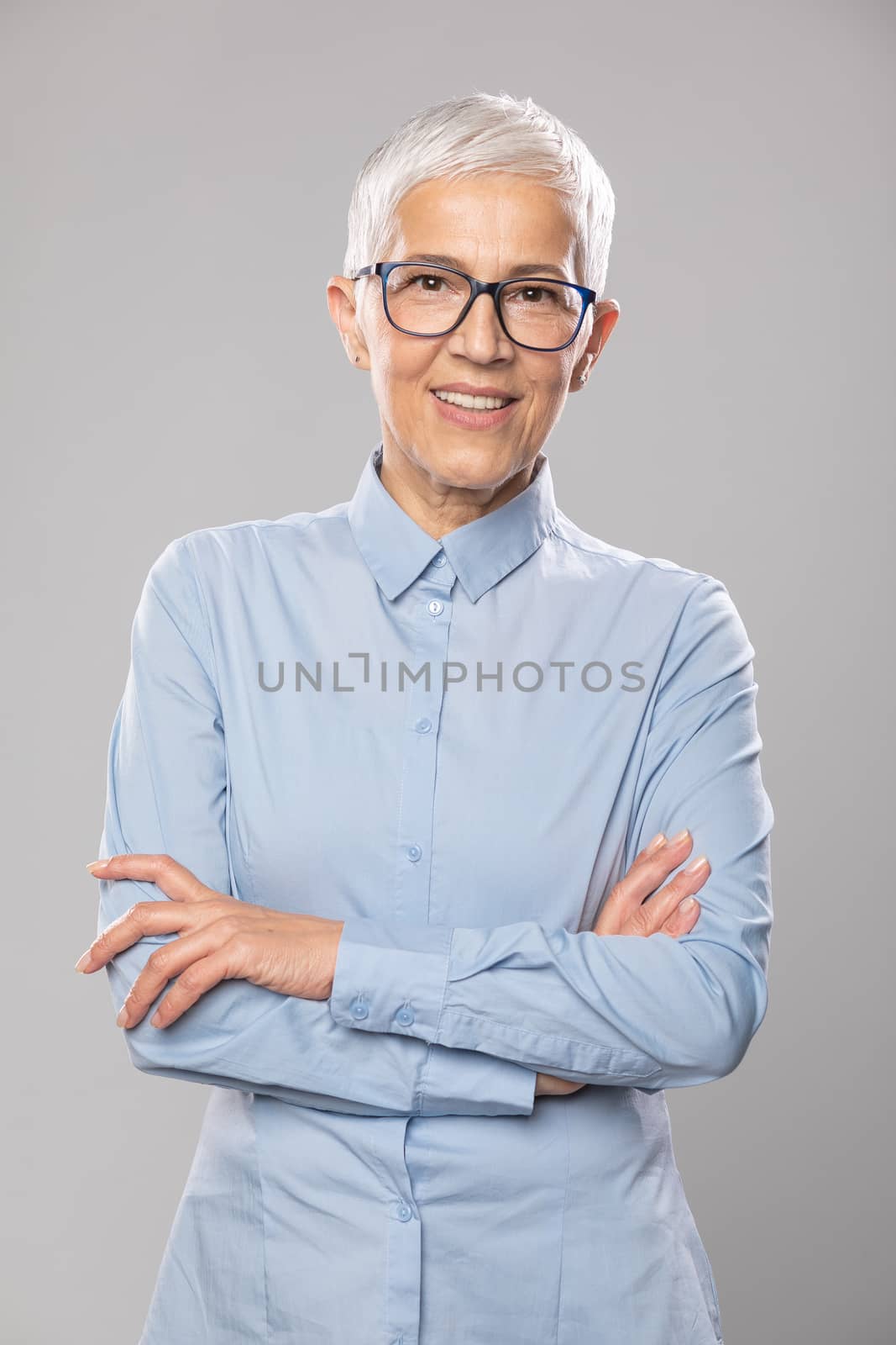 Beautiful smiling cute senior businesswoman with glasses and a blue shirt with short white hair and glasses posing in front of gray background