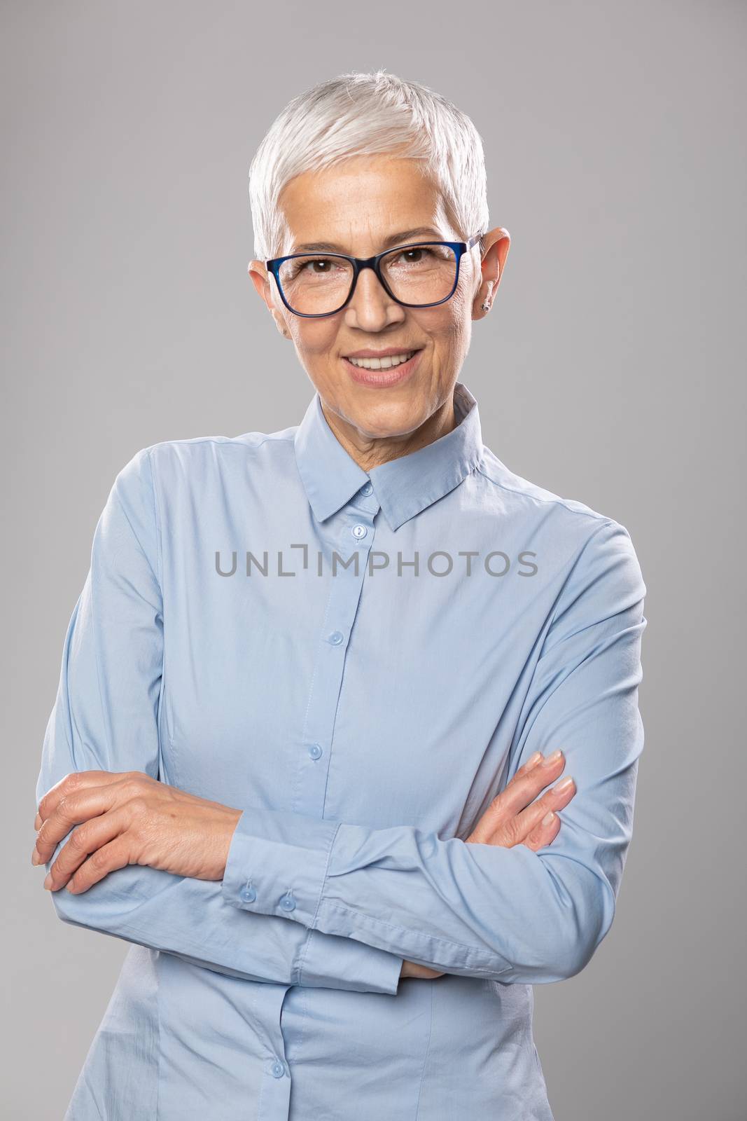 Beautiful smiling cute senior businesswoman with glasses and a blue shirt with short gray white hair and glasses posing in front of gray background