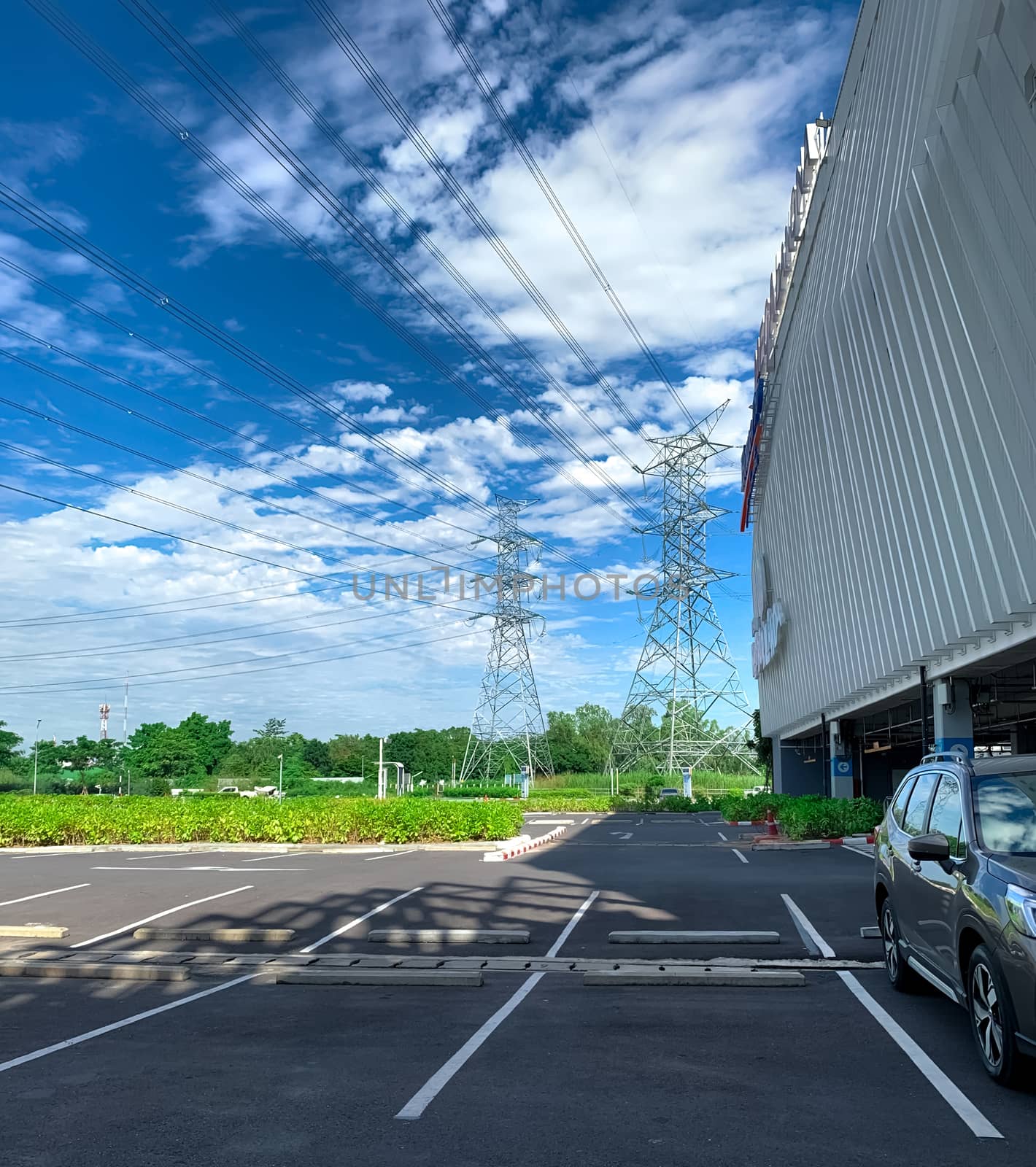 Car parking space at shopping mall for customer service. Outdoors asphalt car parking lot on sunny day with green tree forest and high voltage electric tower. Overhead transmission line. 