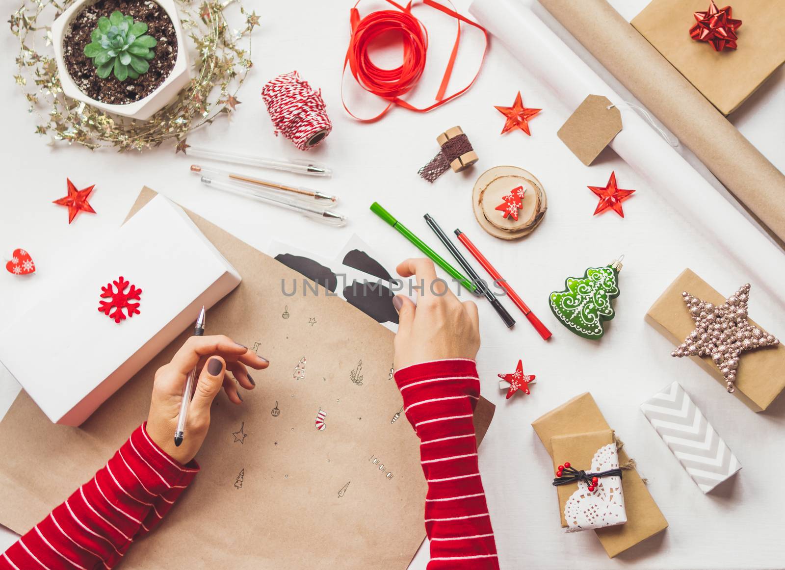 Top view on table with Christmas decorations. Left handed woman draws New Year symbols on craft paper and wraps presents. Flat lay.