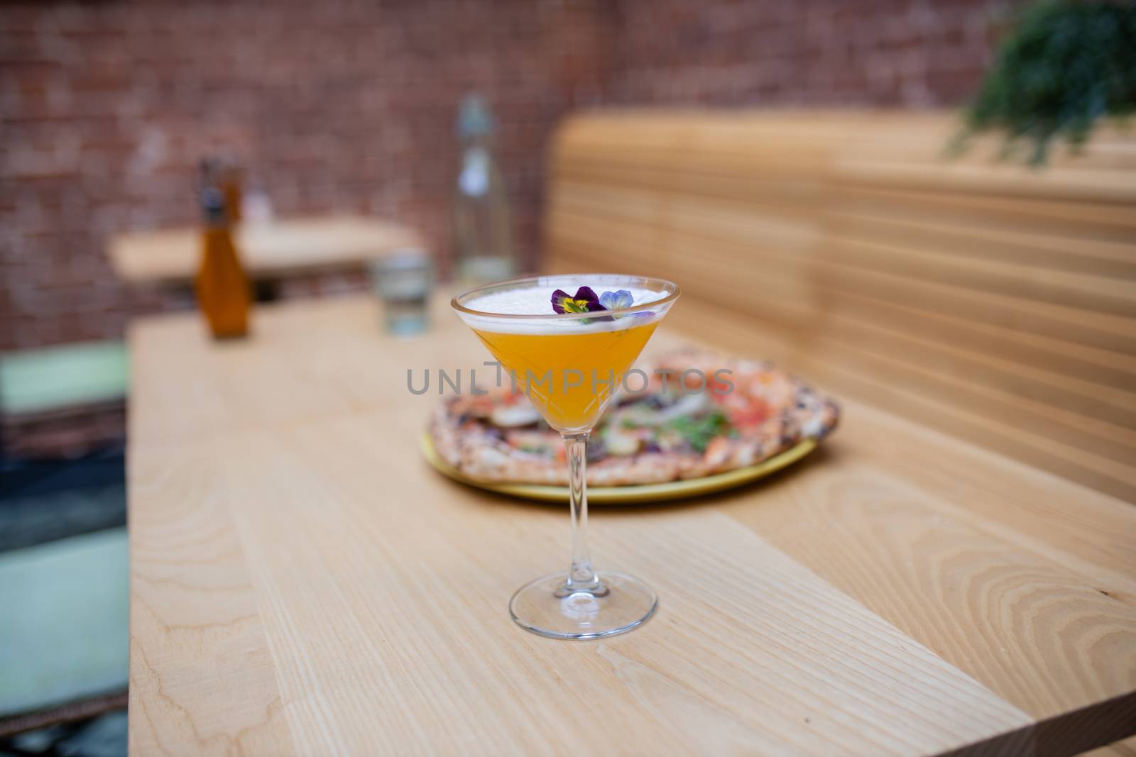 Picture of a yellow cocktail and a thick vegan pizza on a long wooden restaurant table, with a red bricks wall and a long wooden seat as background