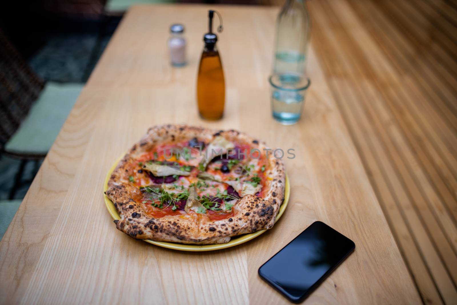 Picture of a tasty looking vegan pizza on a wooden table alongside a bottle of salad dressing, a smartphone, and a blurry glass of mineral water