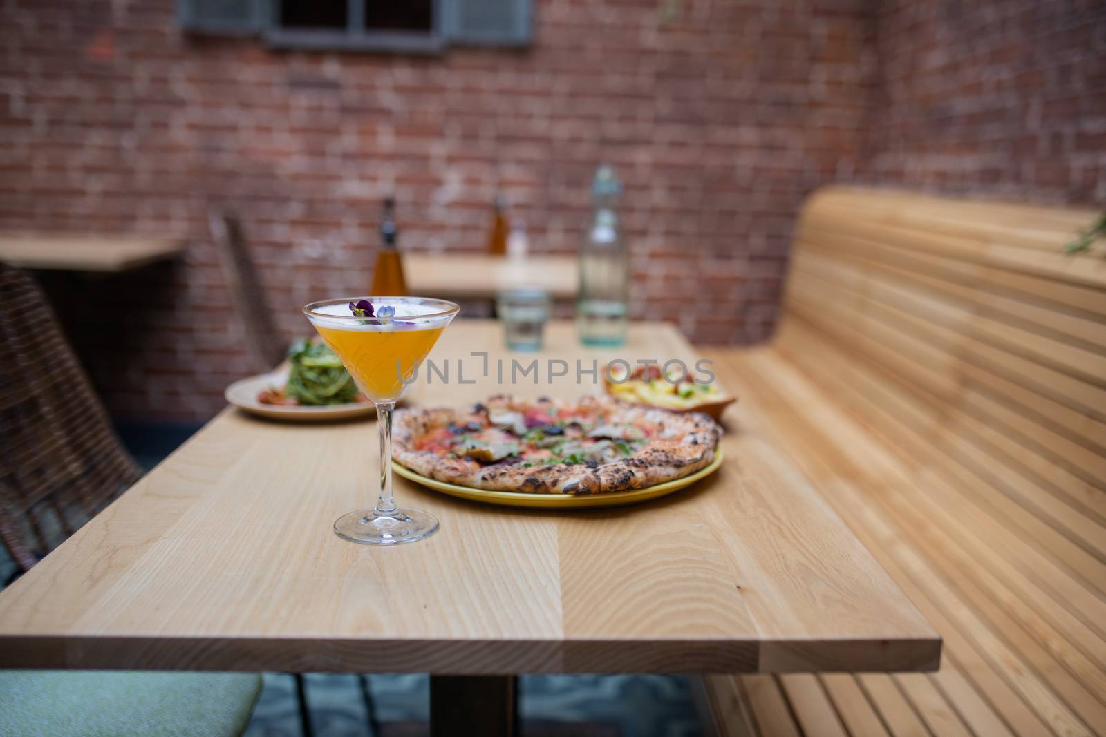 Picture of a vegan pizza and a cocktail on a wooden restaurant table, with french air fries, zucchini noodles, and a salad dressing bottle on the same table and with a brick wall as background