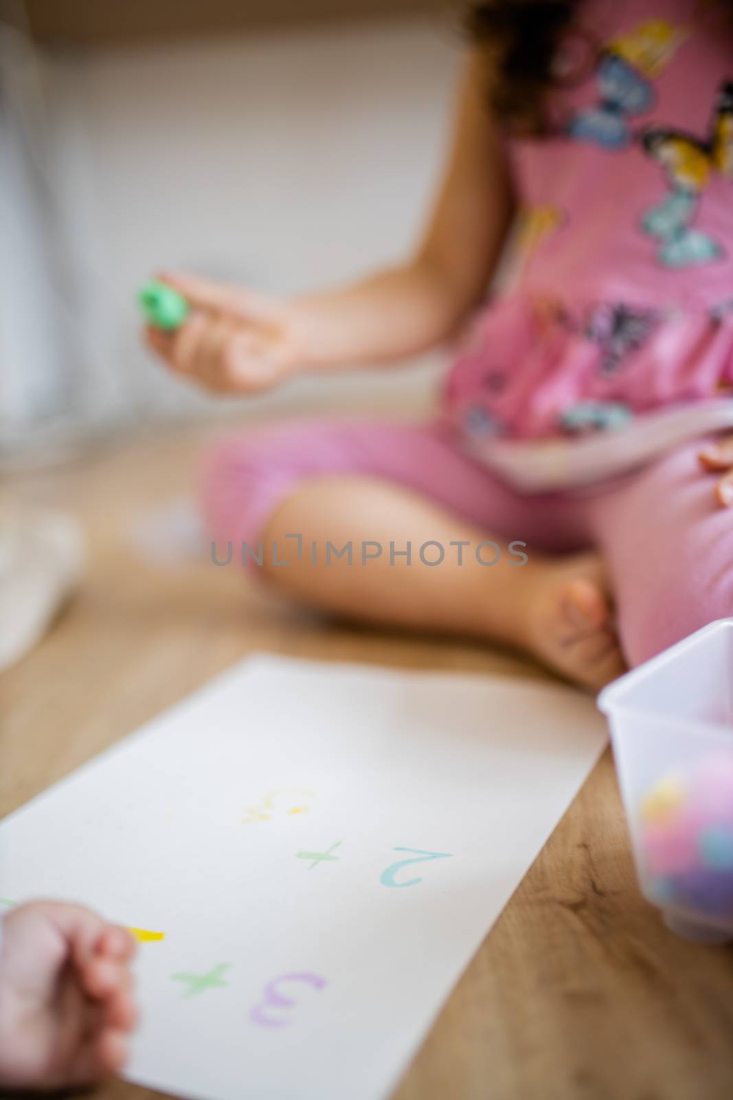 Portrait picture of a little girl in a pink dress sitting on the wooden floor and doing sums on a paper sheet with a green textmarker, alongside a plastic box and blurry baby hand