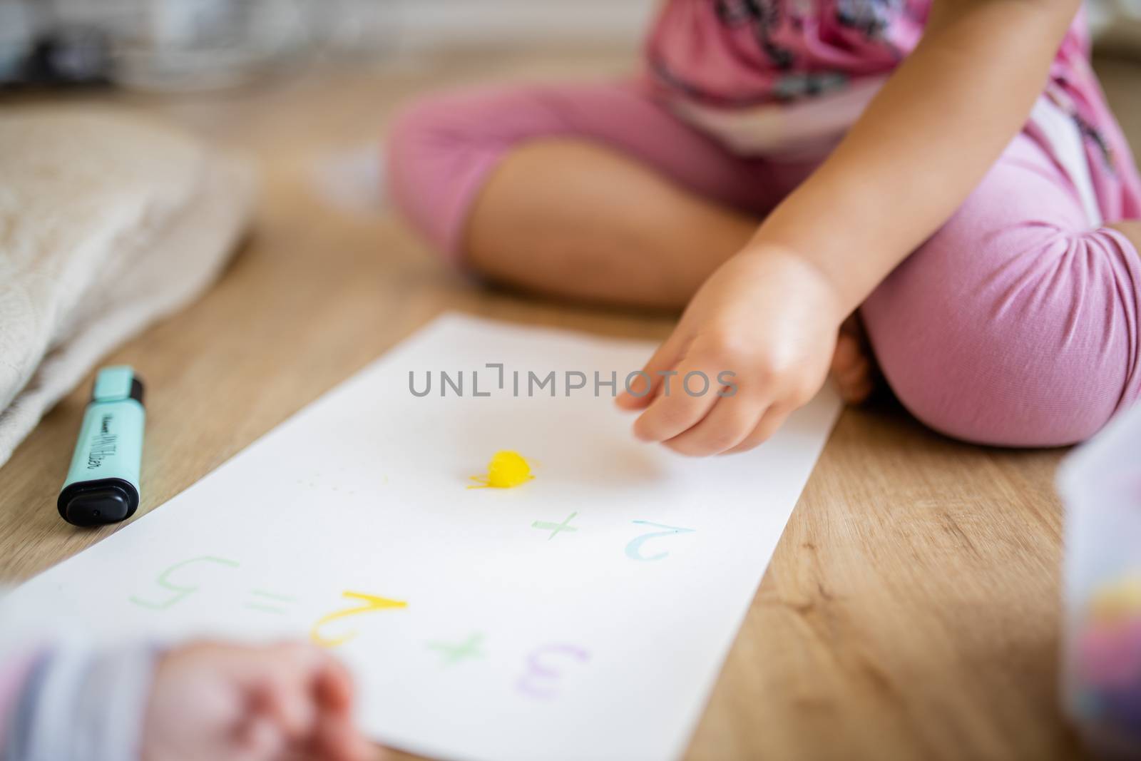 Picture of the legs of a little girl sitting on a wooden floor alongside a yellow cotton ball, sums on a paper sheet, and the blurry arm of a baby