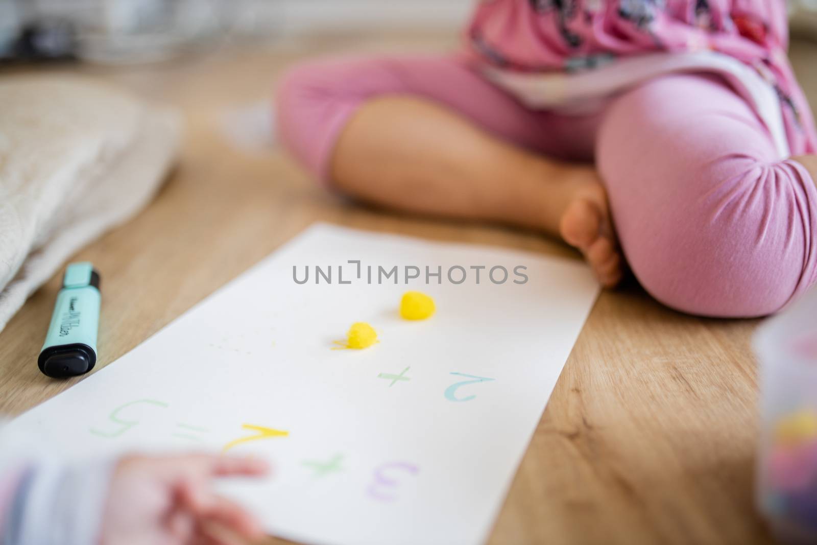 Picture of the legs of a little girl sitting on a wooden floor alongside two yellow cotton balls, sums on a paper sheet, and the blurry arm of a baby