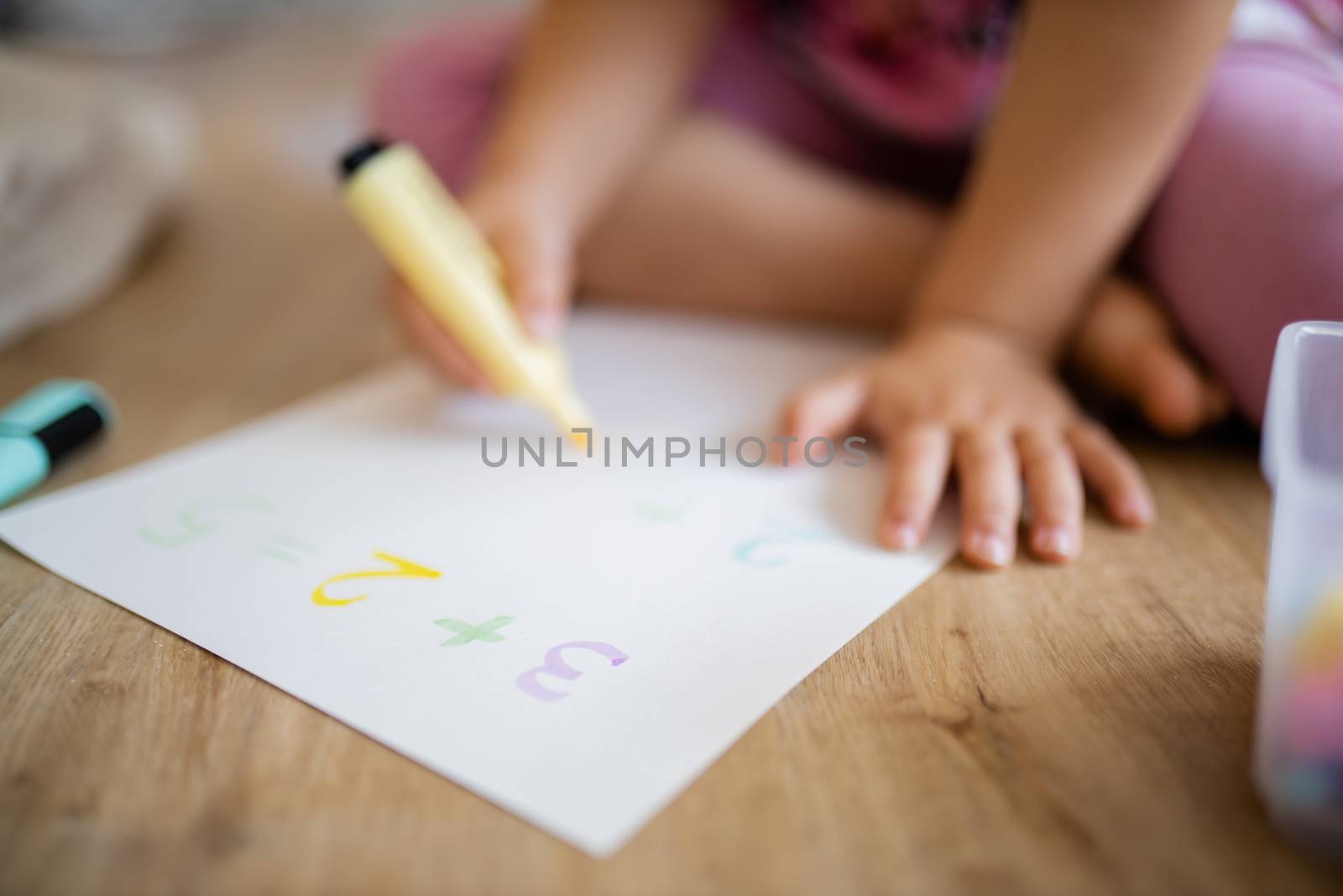 Landscape picture of a little girl in pink clothing sitting on the wooden floor and doing sums on a paper sheet with a yellow textmarker, alongside a plastic box