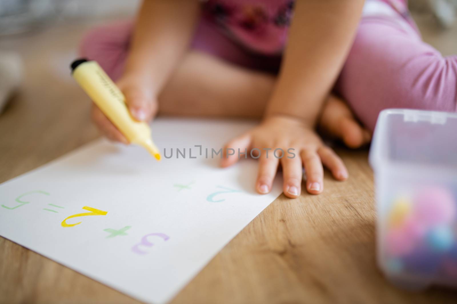 Landscape picture of a little girl in pink clothing sitting on the wooden floor and doing sums on a paper sheet with a yellow textmarker, alongside a plastic box