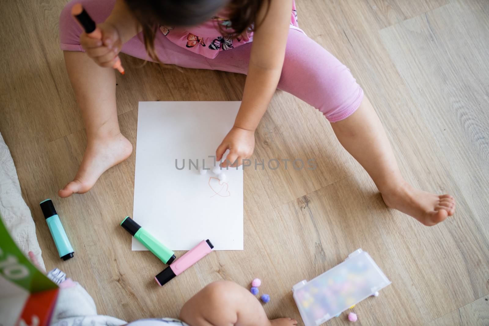 Landscape picture of a little girl in pink clothing sitting on a wooden floor and doing sums on a paper sheet with colorful textmarkers, and cotton balls alongside her baby sibling