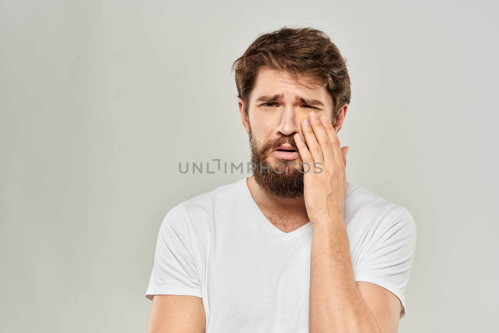 A man in a white t-shirt with a beard emotions displeased facial expression light background by SHOTPRIME
