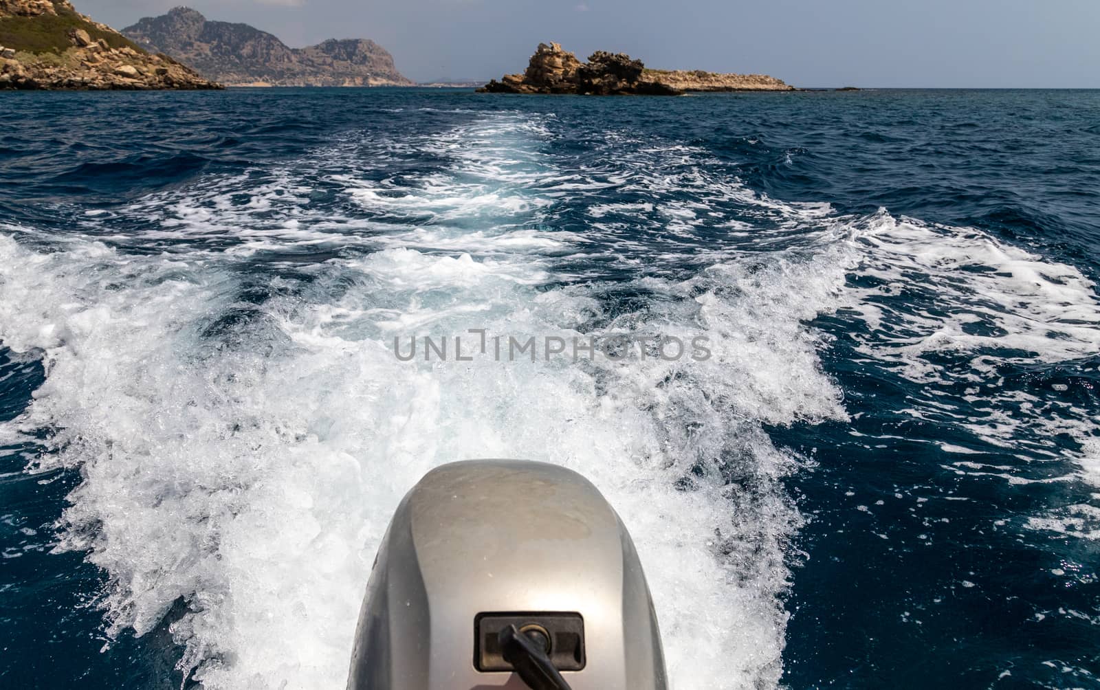 Stern wave of a motor boat with the motor in foreground and the rocky coastline on the eastside of Greek island Rhodes in the background on a sunny day in spring