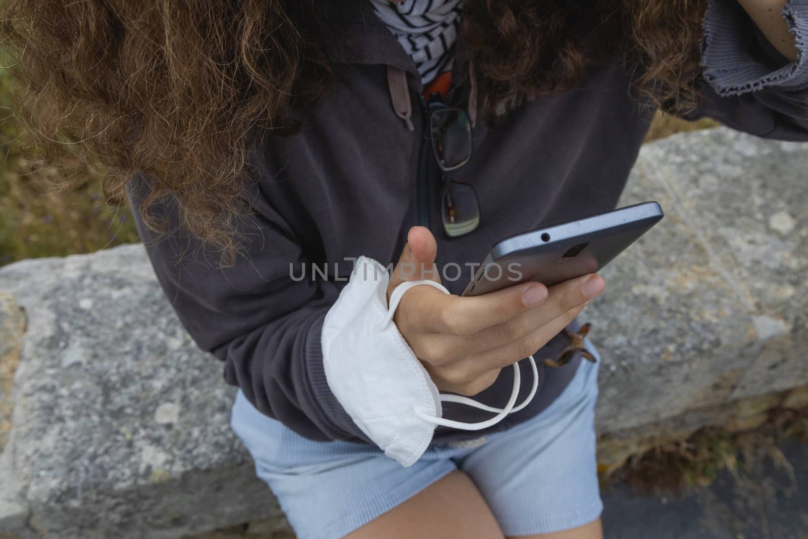 A young woman, grasping a protective face mask with her fingers, holds at the same time, with her hands, a smart mobile phone, Spain.