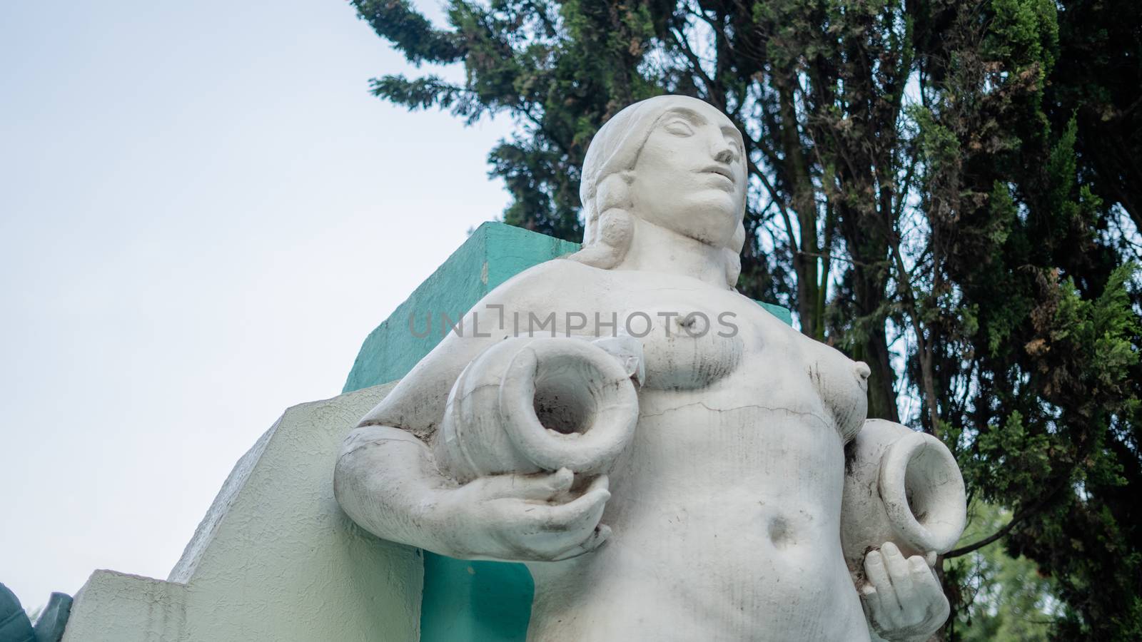 Picture of a naked woman statue holding two vases, with trees as background