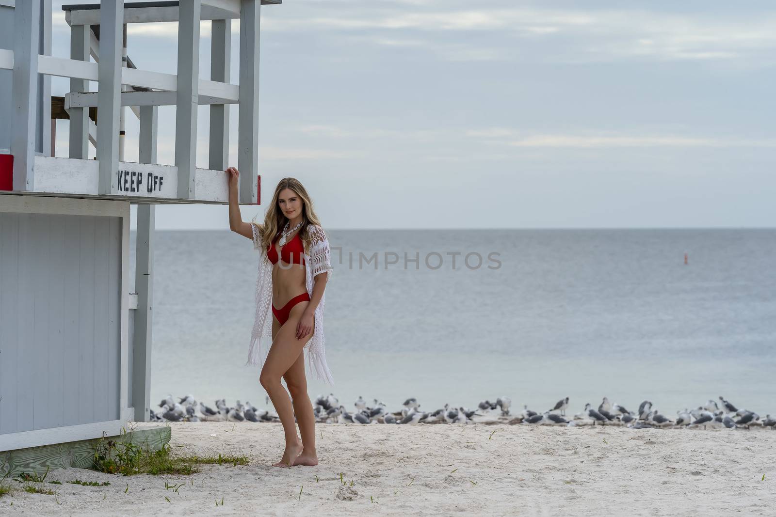 A beautiful blonde bikini model enjoys the weather outdoors on the beach while posing near a lifeguard station