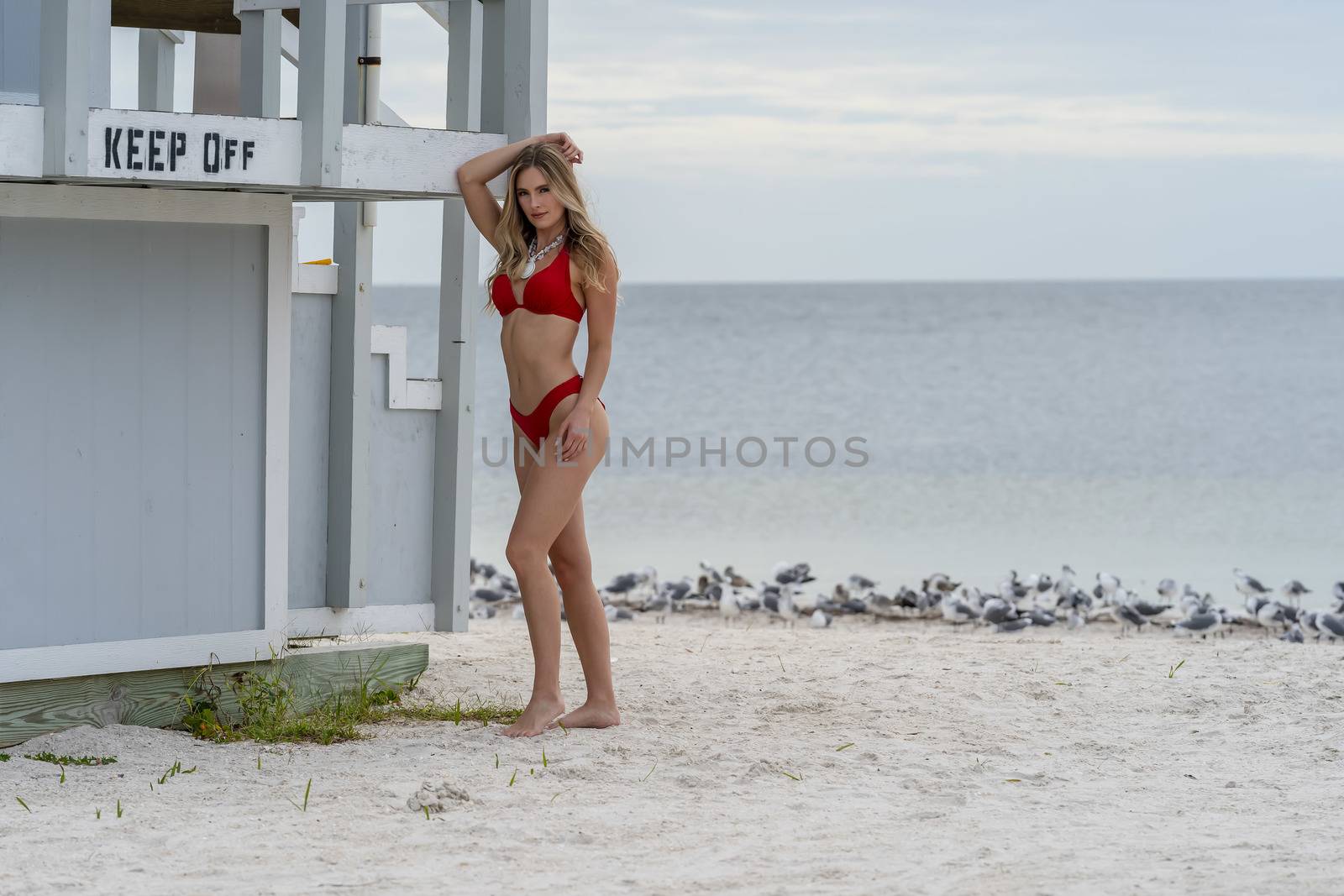 A beautiful blonde bikini model enjoys the weather outdoors on the beach while posing near a lifeguard station