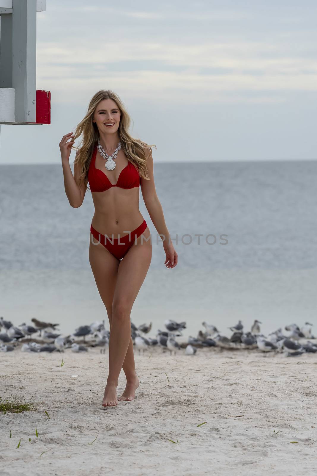 A beautiful blonde bikini model enjoys the weather outdoors on the beach while posing near a lifeguard station