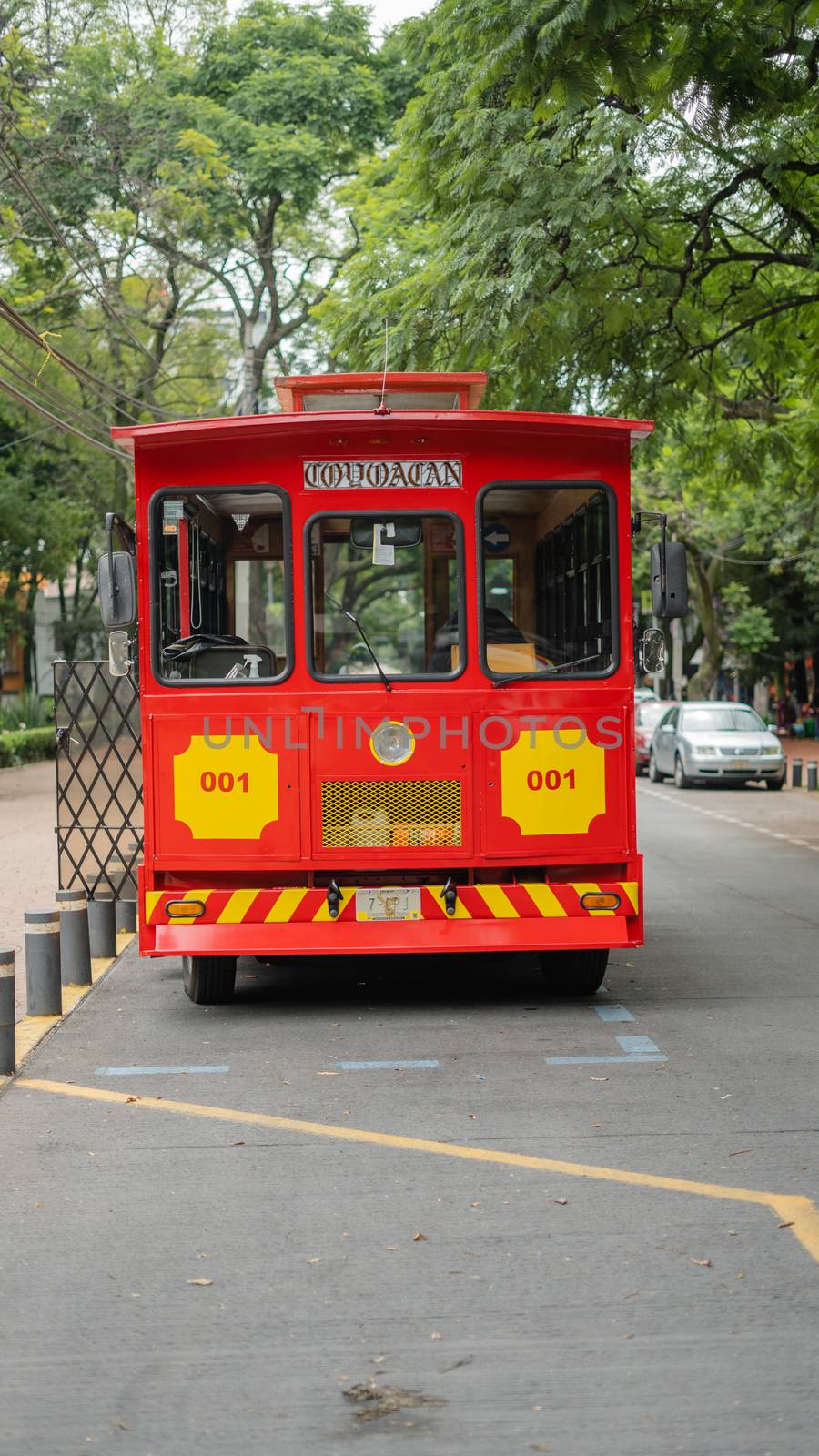 Portrait style picture of a red and yellow tram driving under the trees from Coyoacan